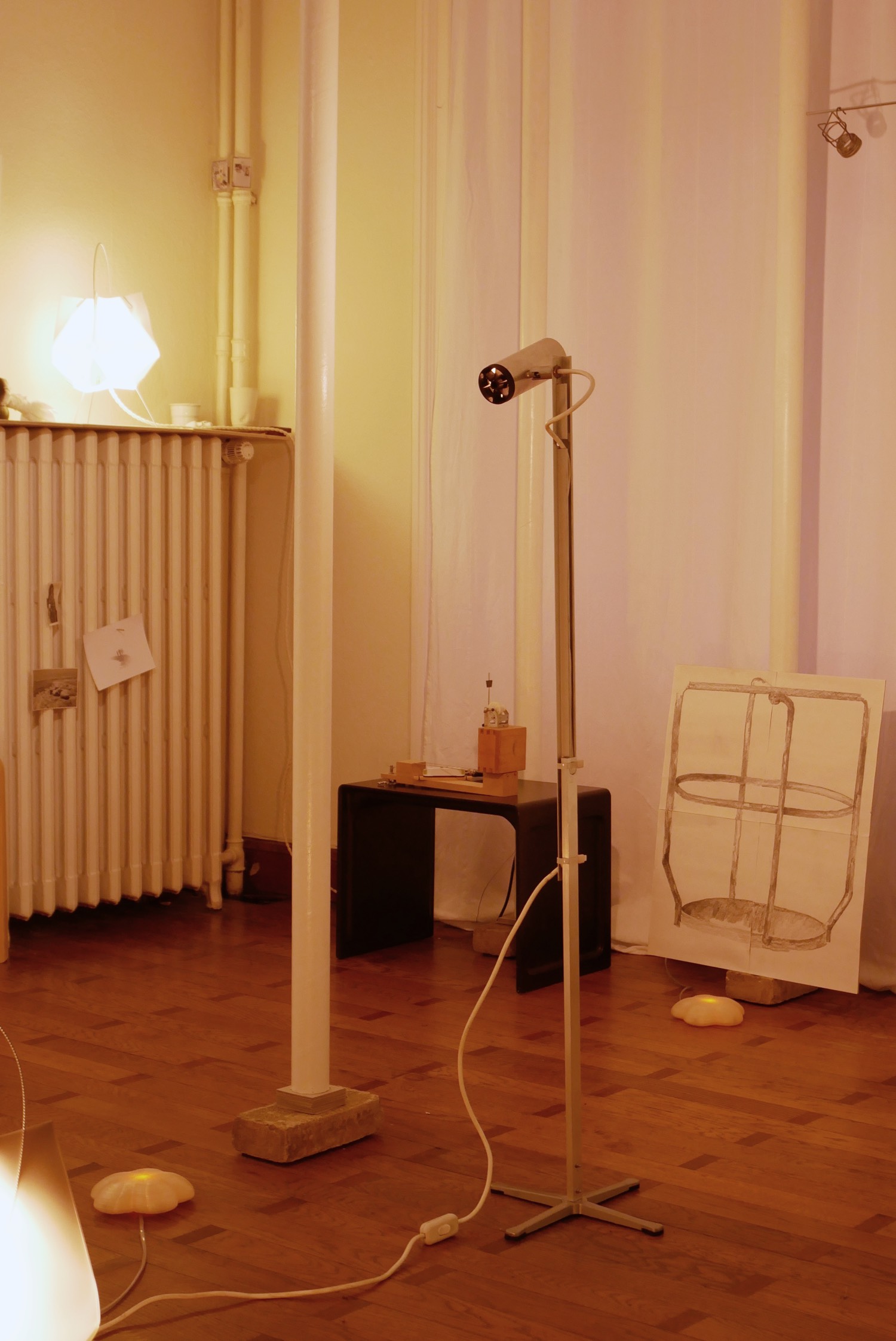 Corner of the room with a musical instrument on a low Vitsoe table, a lamp and cups on a radiator, an industrial-looking floor lamp, a wire lamp socket with a large scale drawing of it below