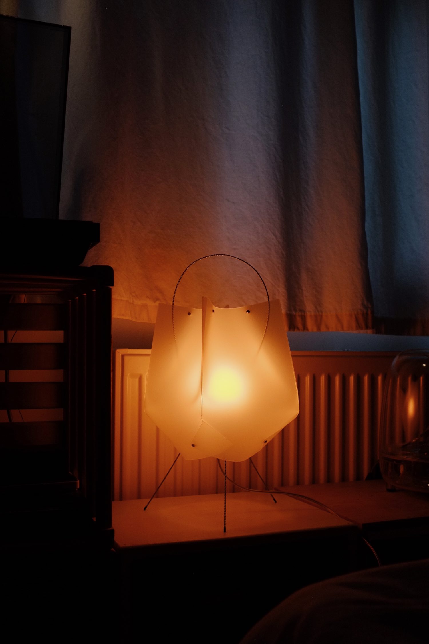 Translucent plastic lamp with wire legs and handle glowing orange on a low side table in front of a white wall and curtain