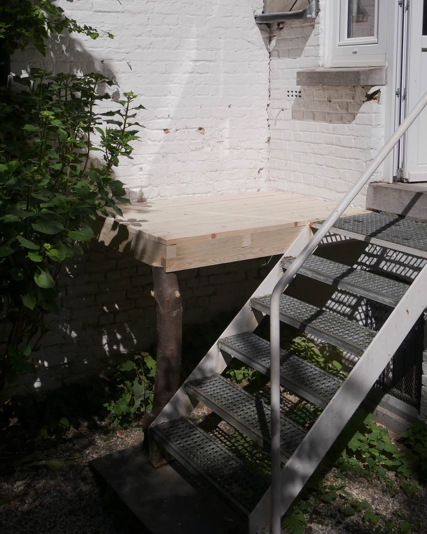 Sun-drenched simple wooden platform in the corner of a leafy courtyard with white brick walls. It is attached to a modernistic metal stair with a white tubular railing