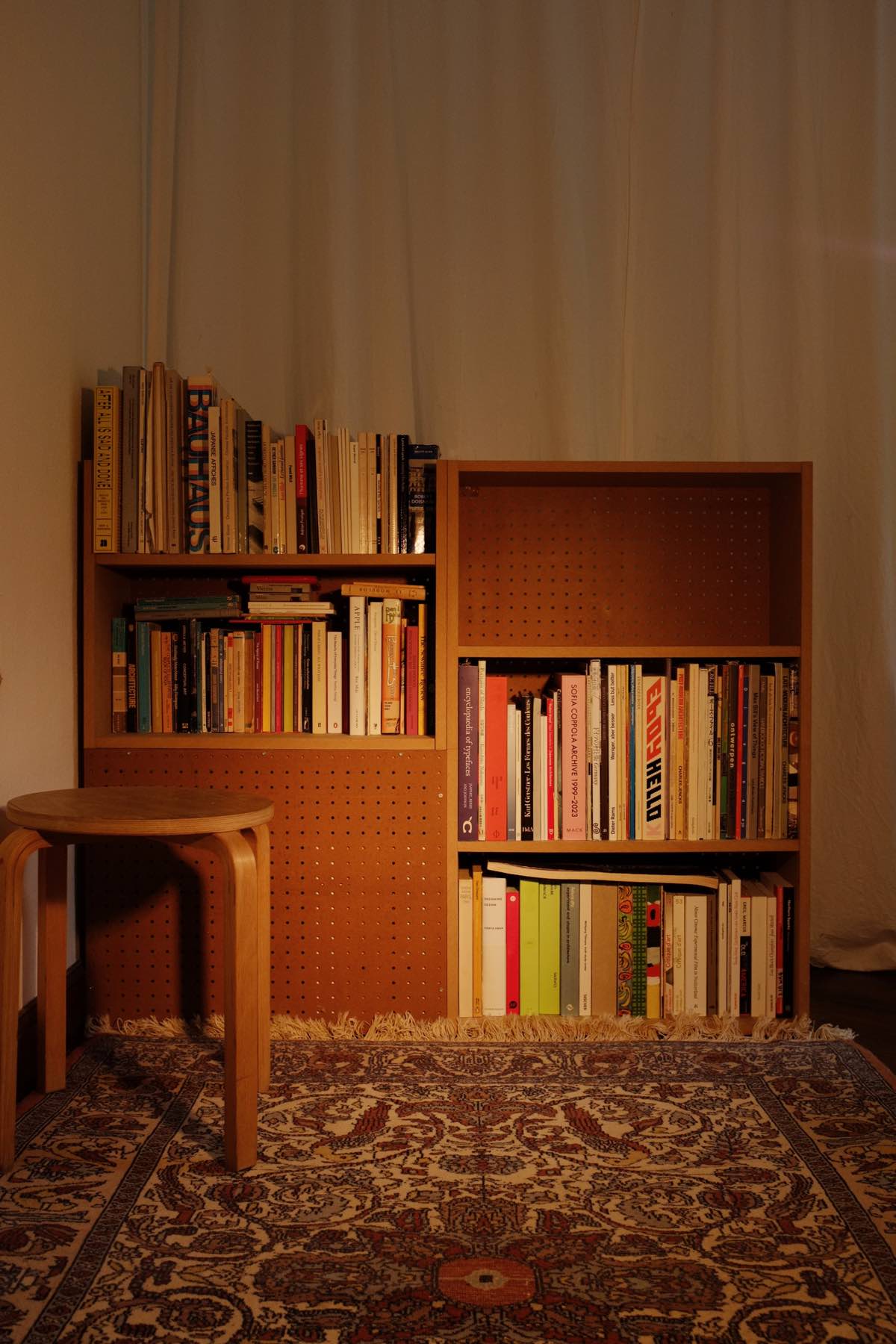 A medium-low bookshelf in a softly lit room. In front is an ornate carpet, and behind is a white curtain. The shelves on the left are offset from those on the right, and the top is open with irregularly sized books sticking out.