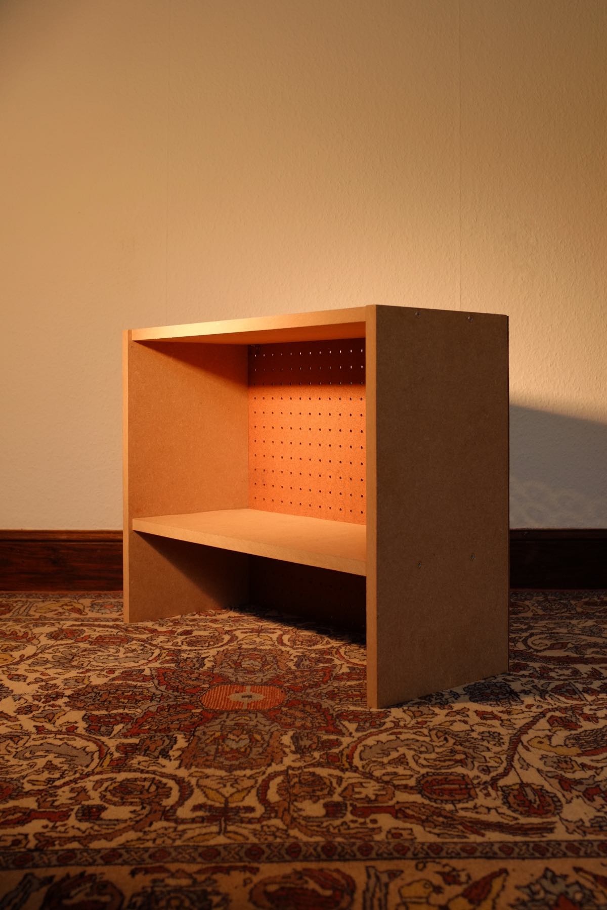 A single unit from the bookshelf above — one enclosed taller compartment above a smaller open compartment. It’s made of dull-coloured MDF with brown pegboard backing.